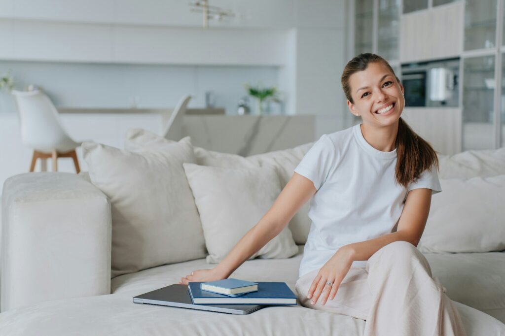 Purposeful woman in casual clothes sits on couch with laptop and books, happy after remote lesson